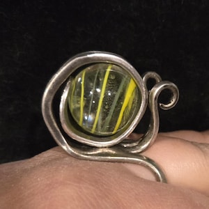 May include: A unique silver ring featuring a clear glass marble with yellow and white stripes. The ring has a swirling, artistic design around the marble, with a decorative flourish on the side. The ring is shown on a person's finger.