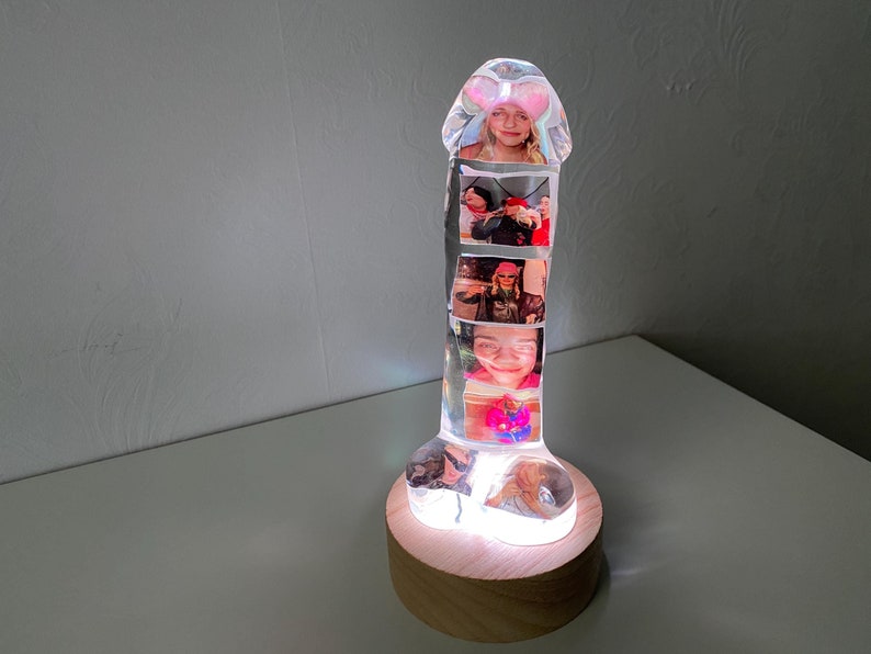Personalized phallus-shaped resin lamp with embedded photos arranged vertically inside the clear resin. The lamp sits on a wooden base with LED lighting, highlighting the vibrant colors of the photos.