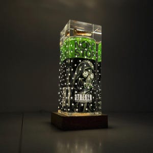 Puede incluir: Una pantalla iluminada con un objeto cilíndrico con el texto "NON STOP" y "STALKER". El objeto está encerrado en una caja de acrílico transparente con pequeñas luces. La base es un bloque de madera oscura.