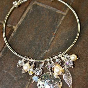 May include: Silver-toned charm bracelet with various dangling charms. Charms include pearls, crystals, and a silver disc engraved with "Always in my Heart." The bracelet is displayed on a dark, textured surface.