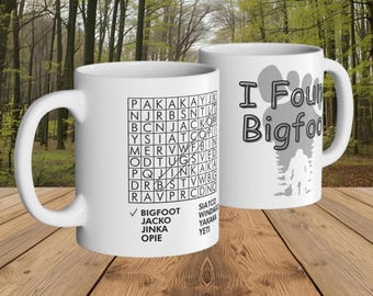 May include: Two white ceramic mugs with black text and graphics. One mug features a Bigfoot word search puzzle. The other mug has the text "I Found Bigfoot" with a Bigfoot silhouette. The mugs are on a wooden surface.
