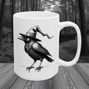 May include: White ceramic mug featuring a black and white illustration of a raven wearing a witch's hat. The raven is standing with its claws visible. The mug has a large handle and is set against a blurred forest background.