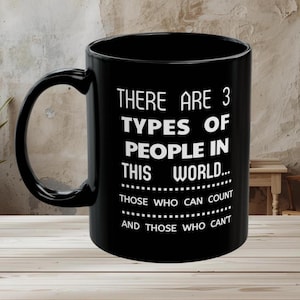 Puede incluir: Taza de cerámica negra con una cita humorística blanca: "THERE ARE 3 TYPES OF PEOPLE IN THIS WORLD... THOSE WHO CAN COUNT AND THOSE WHO CAN'T." La taza tiene un asa grande y está sobre una superficie de madera.