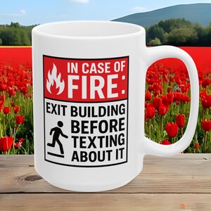 May include: White ceramic mug with a black and red graphic that reads "IN CASE OF FIRE: EXIT BUILDING BEFORE TEXTING ABOUT IT." The mug has a large handle and is set against a background of red flowers.