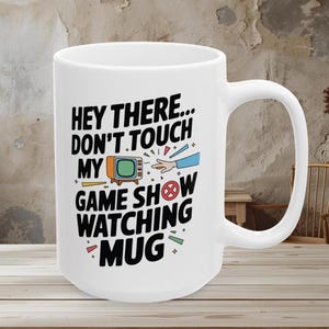 May include: White ceramic mug with a large handle. The mug features the text "HEY THERE... DON'T TOUCH MY GAME SHOW WATCHING MUG" in black, with a retro TV graphic and colorful accents. A fun gift for TV lovers.