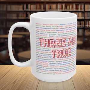 May include: White ceramic mug with the words "THREE ARE TRUE" in red and blue. The mug is covered in a list of words in various colors. The mug has a large handle and is set against a blurred background of bookshelves.