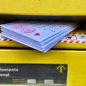 May include: A yellow metal mailbox with a red and white striped border. The mailbox has a slot for mail and a sign that says "d&eacute;partements international". The mailbox is labeled "La Poste" and has a sign that says "Derni&egrave;re lev&eacute;e lundi au vendredi 17H00 samedi 12H00".