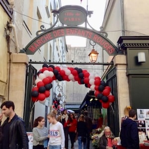 May include: The entrance to the Marche des Enfants Rouges in Paris features a dark metal archway with red lettering and a large balloon arch in red, white, and black colors. The archway is topped with a sign reading Ville de Paris.
