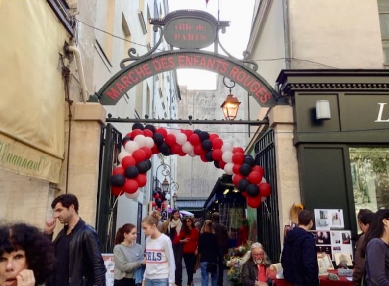 May include: The entrance to the Marche des Enfants Rouges in Paris features a dark metal archway with red lettering and a large balloon arch in red, white, and black colors. The archway is topped with a sign reading Ville de Paris.