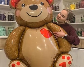 Può includere: Un grande palloncino a forma di orsetto marrone con dettagli rossi sulle orecchie, sulle zampe e un cuore sul petto. Il palloncino ha una faccia sorridente ed è tenuto da una persona.