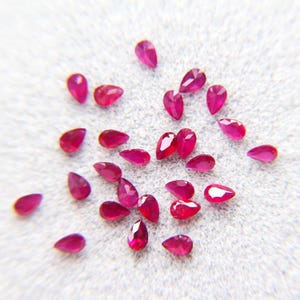 May include: A collection of small, pear-shaped, faceted rubies. The gemstones are a vibrant, deep red color and are scattered across a light gray surface. The facets catch the light, creating a sparkling effect.