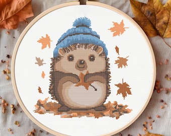 May include: A cute cartoon hedgehog wearing a blue hat and holding a brown leaf. The hedgehog is surrounded by brown leaves on a white background.