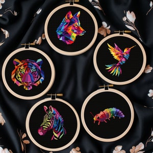 May include: Five cross-stitch embroidery hoops featuring geometric animal designs. The animals include a wolf, tiger, hummingbird, zebra, and a caterpillar, all rendered in vibrant rainbow colors against a black background. The hoops are made of wood.