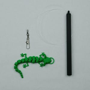 May include: A green lizard-shaped toy with a swiveling head and a black cord. The toy is attached to a black cord with a silver swivel clip.
