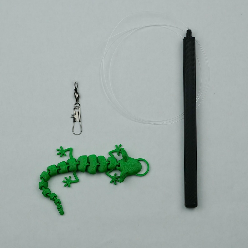 May include: A green lizard-shaped toy with a swiveling head and a black cord. The toy is attached to a black cord with a silver swivel clip.