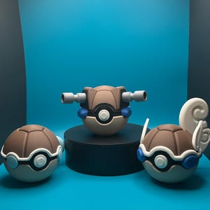 May include: Three 3D printed Pokemon figures, all brown with white and blue accents. The centre figure has two grey cannons on the top. The figures are on a blue surface.