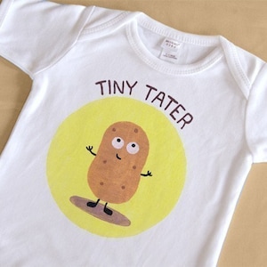Może przedstawiać: Biały śpioszek dla niemowlęcia z żółtym kółkiem i kreskówkową postacią ziemniaka z oczami i uśmiechem. Tekst "Tiny Tater" jest napisany nad ziemniakiem.