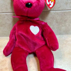 May include: A pink Beanie Baby plush toy with a white heart on its chest. The toy has a red Ty tag with a white star and the word "Ty" in white.