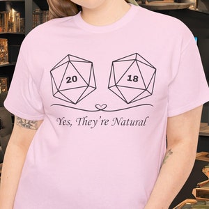 Puede incluir: Camiseta rosa claro con dos dados que muestran los números 20 y 18, y el texto "Yes, They're Natural". La persona lleva vaqueros azules.