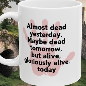 Könnte beinhalten: Weiße Keramik-Kaffeetasse mit einem schwarzen und rosafarbenen Handabdruck-Design. Die Tasse trägt den Text "Almost dead yesterday, Maybe dead tomorrow, but alive, gloriously alive, today".