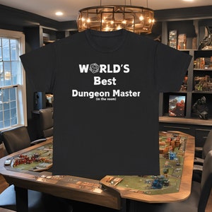 Puede incluir: Camiseta negra con texto blanco que dice "WORLD'S Best Dungeon Master (in the room)" y una imagen blanca de un dado de 20 caras.