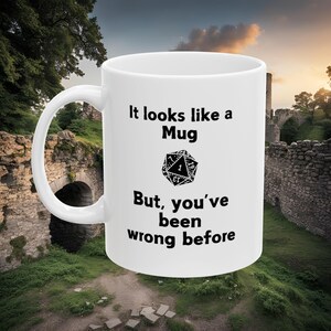 Op de afbeelding: Een witte keramische mok met een zwarte afbeelding van een 20-zijdige dobbelsteen en de tekst "It looks like a Mug But, you've been wrong before".
