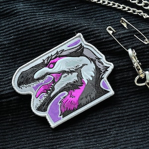 May include: A white and purple embroidered patch featuring a detailed illustration of a dinosaur head with sharp teeth and a fierce expression.