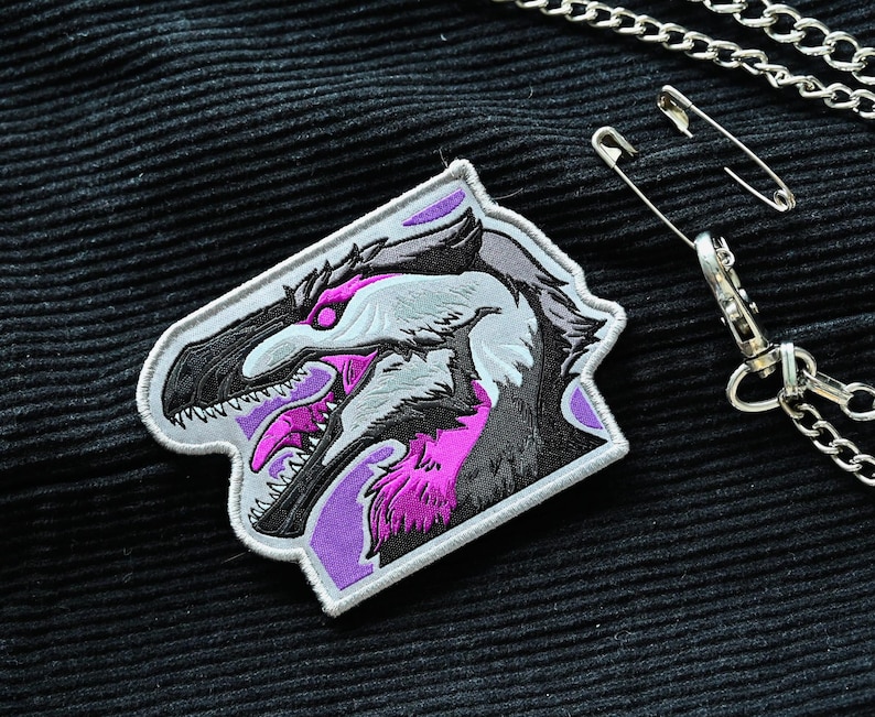 May include: A white and purple embroidered patch featuring a detailed illustration of a dinosaur head with sharp teeth and a fierce expression.