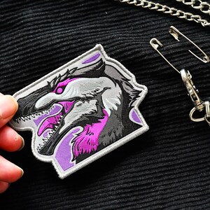 May include: A purple and gray embroidered patch featuring a detailed illustration of a dinosaur head with sharp teeth.