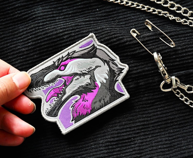 May include: A purple and gray embroidered patch featuring a detailed illustration of a dinosaur head with sharp teeth.