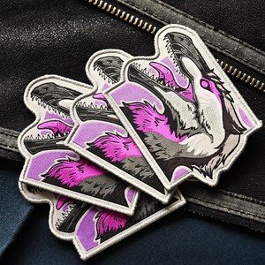 May include: Set of four embroidered patches featuring a pink and gray wolf with a purple background. The wolf's mouth is open and its teeth are visible. The patches are sewn onto a black leather jacket.