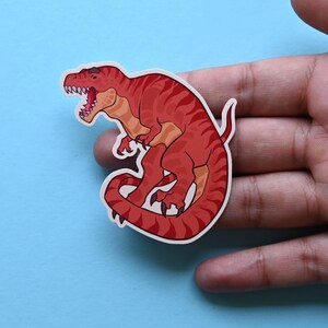 May include: Red dinosaur sticker with a curled tail and open mouth. The dinosaur is facing left and has a white outline.
