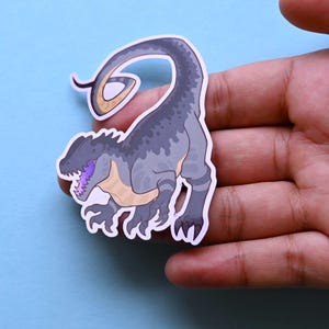 May include: A gray and black dinosaur sticker with a purple tongue and a curled tail. The sticker is on a blue background.