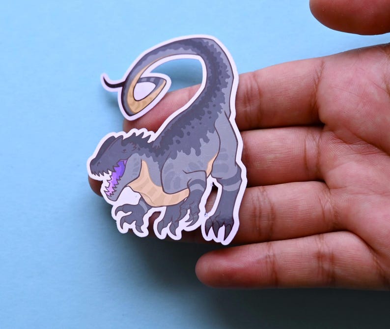 May include: A gray and black dinosaur sticker with a purple tongue and a curled tail. The sticker is on a blue background.