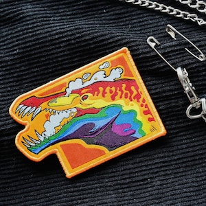 Pode incluir: Um remendo bordado com cores do arco-íris que apresenta um monstro de desenho animado com um sorriso largo e uma nuvem de fumaça. O remendo está preso a uma corrente prateada com um alfinete de segurança.