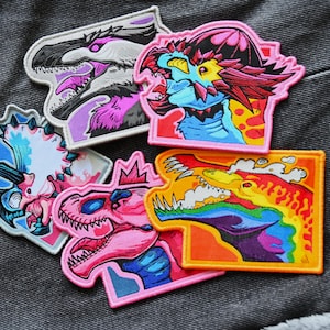 以下が含まれることがあります： 異なる恐竜のデザインが施された、カラフルな刺繍パッチが5つ。パッチは濃いグレーのデニム生地の上に置かれています。恐竜は、ピンクとブルーのティラノサウルス、レインボーのたてがみを持つイエローとブルーの恐竜、レインボーのしっぽを持つブルーとイエローの恐竜、ピンクとブルーのトリケラトプス、パープルのたてがみを持つホワイトとピンクの恐竜です。