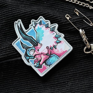 Pode incluir: Um remendo bordado com um triceratops estilizado com um chifre branco e uma gola preta. O remendo é rosa e azul e tem uma borda branca.