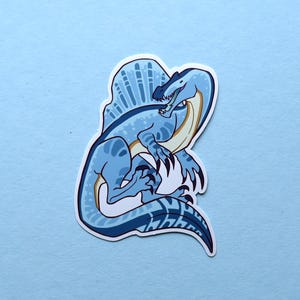 May include: A blue and white cartoon illustration of a Spinosaurus dinosaur with a large sail on its back. The dinosaur is depicted in a playful pose with its mouth open and its claws extended.