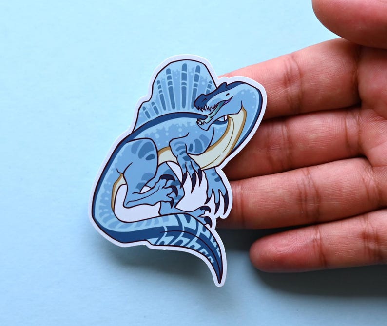 May include: A blue and white cartoon illustration of a Spinosaurus dinosaur with a large sail on its back. The dinosaur is depicted in a playful pose, with its mouth open and its tongue sticking out.