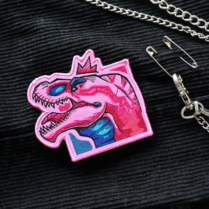 Puede incluir: Parche bordado rosa con un dinosaurio con una corona en la cabeza. El dinosaurio está en un marco cuadrado con un fondo azul y rosa.