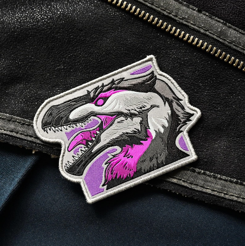 May include: A purple and white embroidered patch featuring a detailed illustration of a dinosaur head with sharp teeth and a pink tongue.