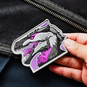May include: A purple and gray embroidered patch featuring a dinosaur head with sharp teeth and a fierce expression.