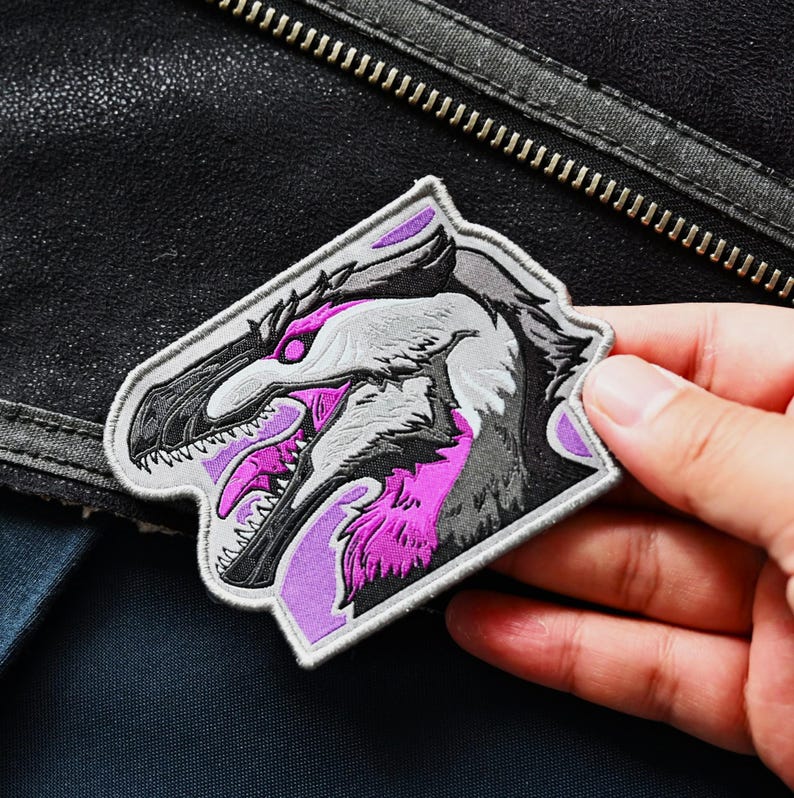 May include: A purple and gray embroidered patch featuring a dinosaur head with sharp teeth and a fierce expression.