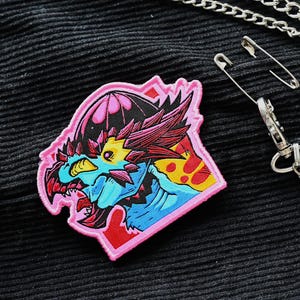 May include: A pink, red, blue and yellow embroidered patch featuring a dragon-like creature with a large head and open mouth. The patch is sewn onto a black fabric background.