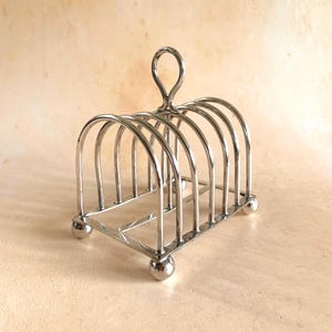 Könnte beinhalten: Ein glänzender, silberfarbener Metall-Toastständer. Der Ständer hat eine rechteckige Basis, die von vier kleinen, runden Füßen getragen wird. Gewölbte Drähte bilden die Schlitze für den Toast, und oben befindet sich ein Schlaufengriff. Er ist für mehrere Toastscheiben konzipiert.