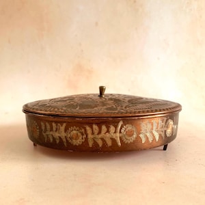 May include: A round copper trinket box with a removable lid featuring an etched floral pattern. The sides of the container are decorated with a repeating silver-toned leaf and flower motif. The box stands on small feet and has a central knob.