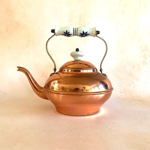 Könnte beinhalten: Eine Kupfer-Teekanne mit einem weiß-blauen floral gemusterten Griff und Deckel. Die Teekanne hat einen gebogenen Ausguss und einen runden Körper. Der Griff ist aus Keramik und Metall. Die Teekanne ist im Vintage-Stil.