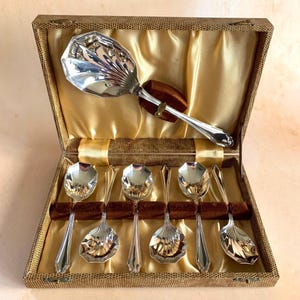May include: A set of silver spoons, including a serving spoon and six smaller spoons, presented in a vintage box. The spoons feature decorative shell-shaped bowls and handles. The box is lined with gold satin and brown velvet.