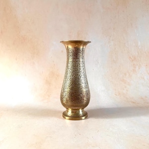 May include: A tall, slender brass vase featuring an intricate, etched floral pattern covering the entire body. The vase has a flared rim, a narrow neck, a bulbous midsection, and a circular pedestal base, all finished in a polished metallic gold tone.