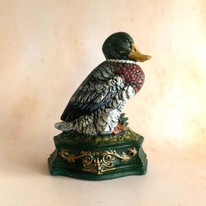 Könnte beinhalten: Detaillierte Entenfigur mit grünem Kopf, gelbem Schnabel und roter und weißer Brust. Die Ente sitzt auf einem grünen Sockel mit goldenen Akzenten. Die Figur ist ein dekorativer Gegenstand.